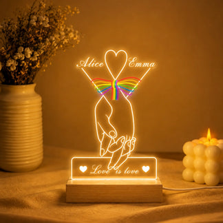 Decorative night light with hands holding a rainbow heart and 'Love is love' text, placed on a surface with a warm glow.