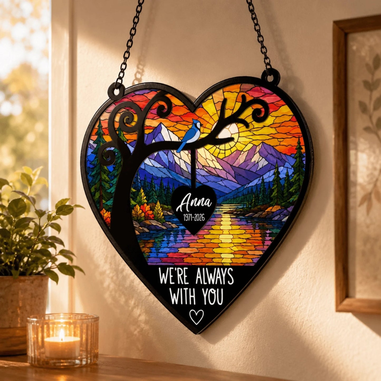 Heart shaped memorial suncatcher with a vibrant stained glass mountain scene and blue jay bird hanging in a window frame.