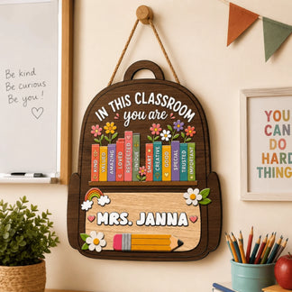 A decorative wooden backpack sign for a classroom with 'In this classroom you are' text, books, and 'Mrs. Janna' name.