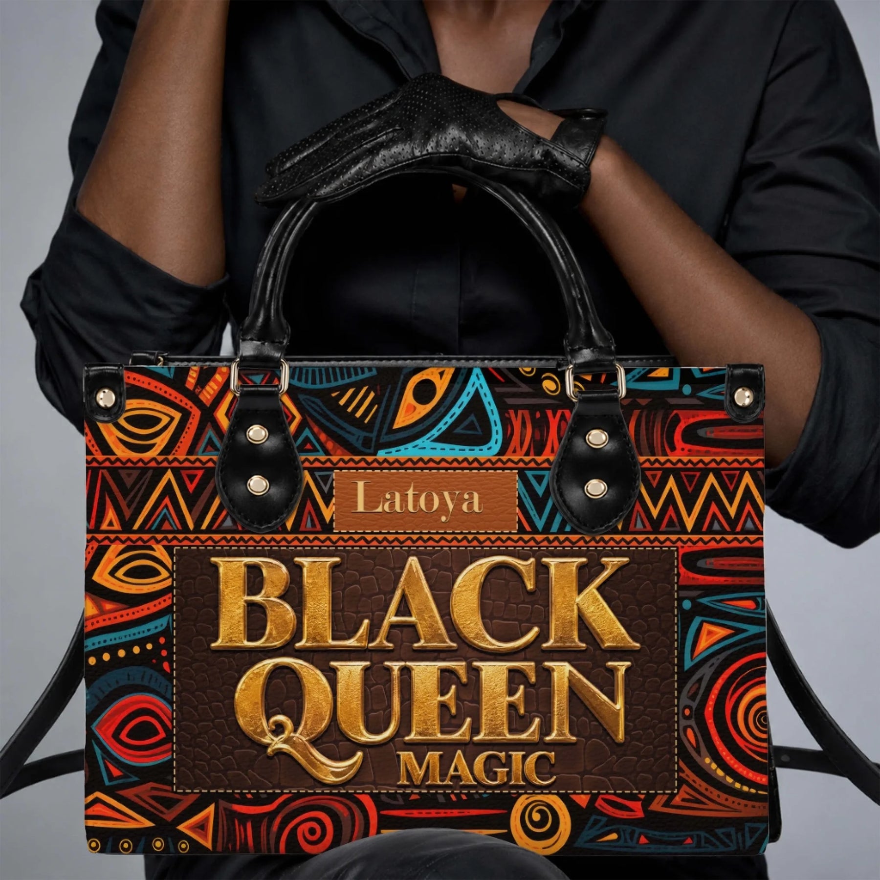 Close up of hands wearing black gloves holding a personalized Black Queen Magic shoulder bag with cultural art motifs.