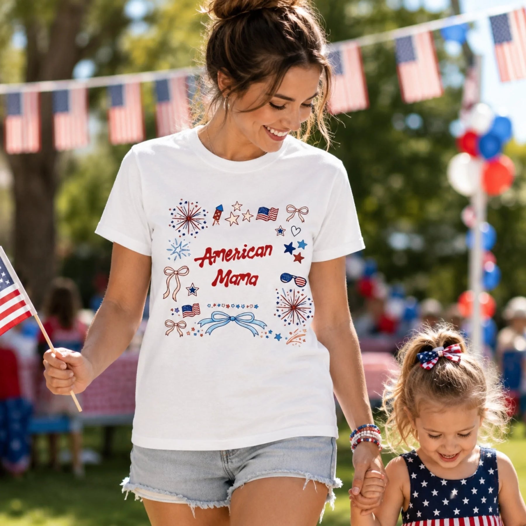 Woman wearing a 'American Mama' t-shirt with a child outdoors, surrounded by American flags and balloons.