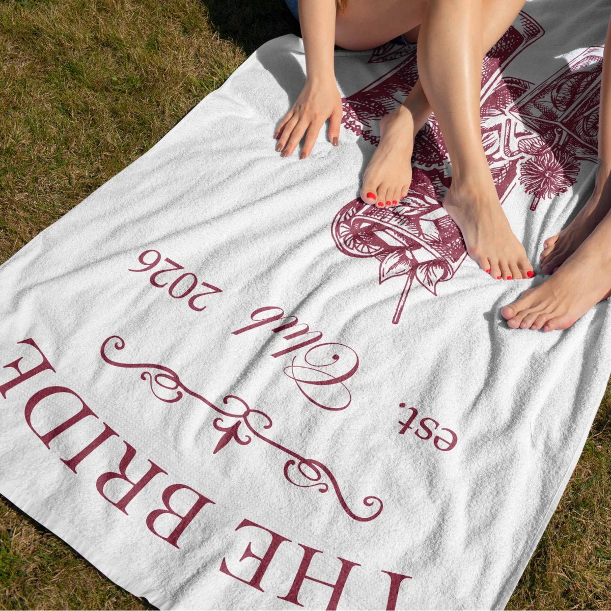 Women relaxing on a custom bridal shower beach towel with cocktail illustrations and The Bride text branding.