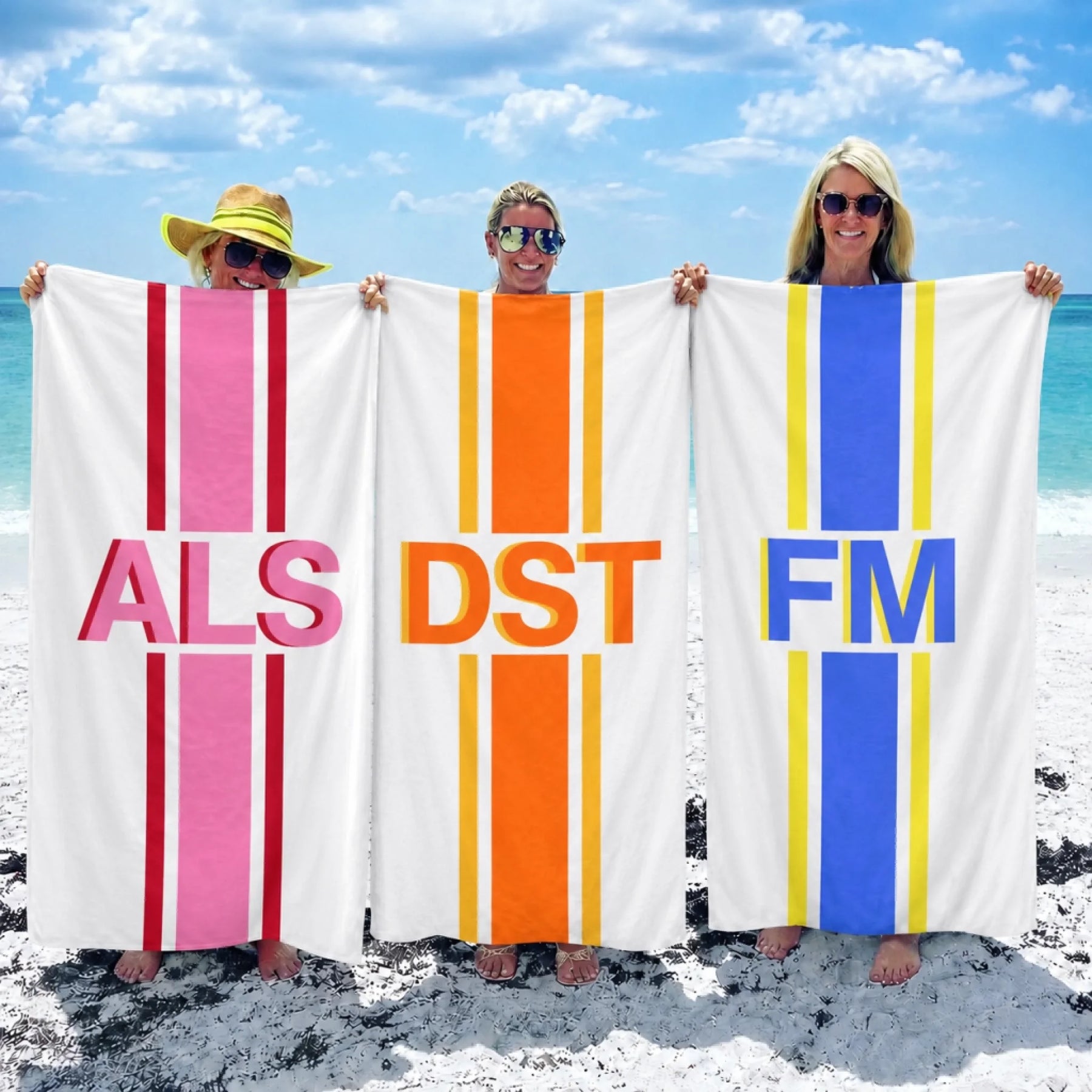 Three women holding custom personalized beach towels with initials ALS DST and FM in vibrant striped designs at beach