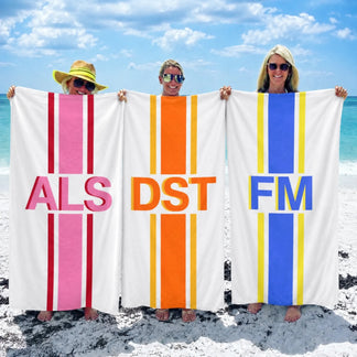 Three women holding custom personalized beach towels with initials ALS DST and FM in vibrant striped designs at beach