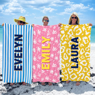 Three women holding custom personalized beach towels with their names printed on blue stripes pink starfish and gold leopard print patterns.