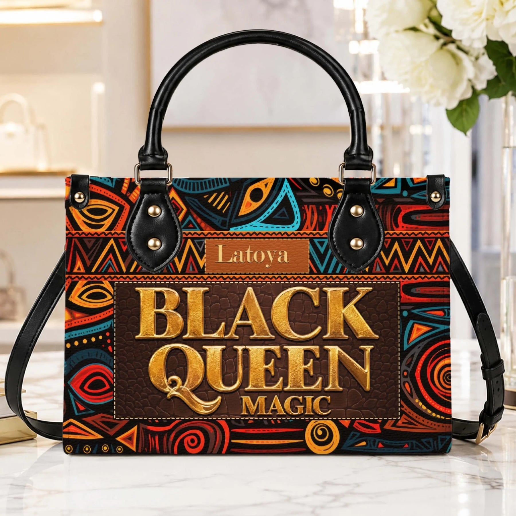 A stylish Black Queen Magic personalized handbag display with a detailed African tribal pattern and gold typography.