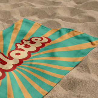 A folded custom beach towel with the name Charlotte and a retro sunburst graphic placed on a sunlit sandy beach.
