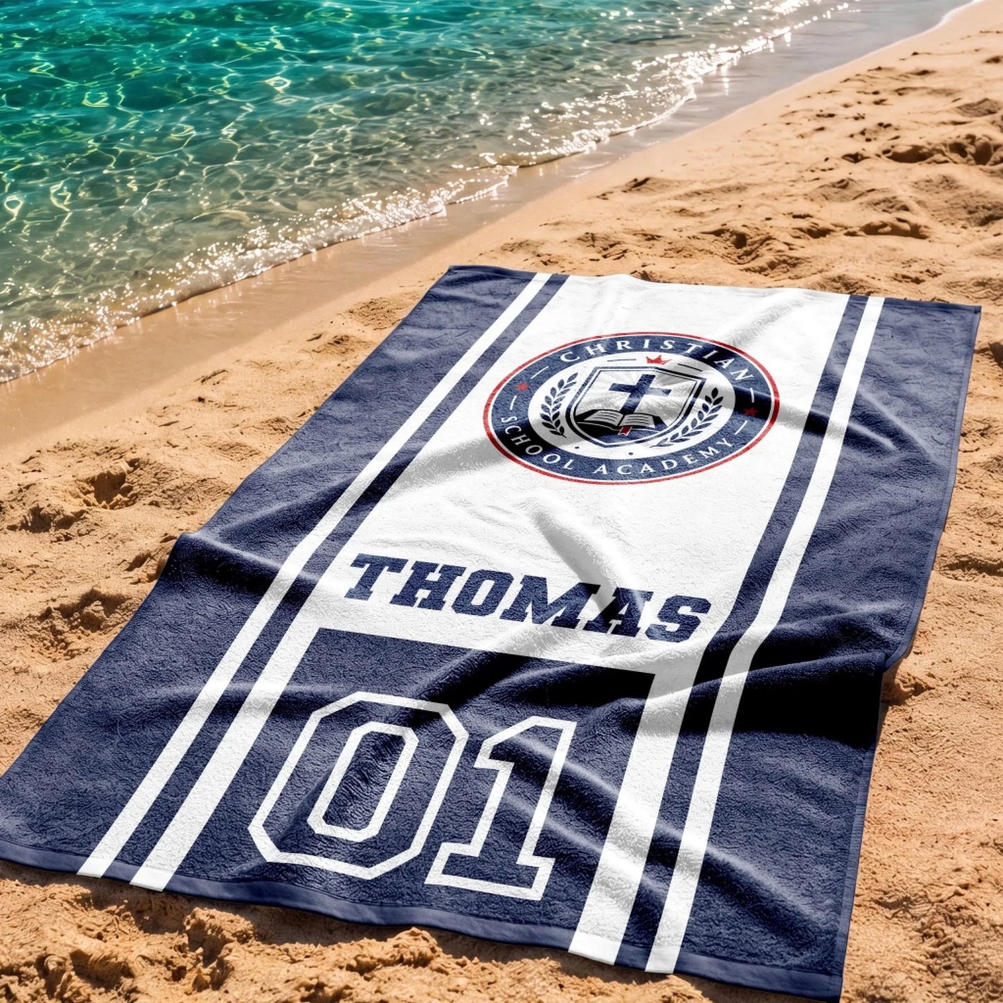 A customized Christian School Academy beach towel featuring the name Thomas and number 01 on the sand by the water.