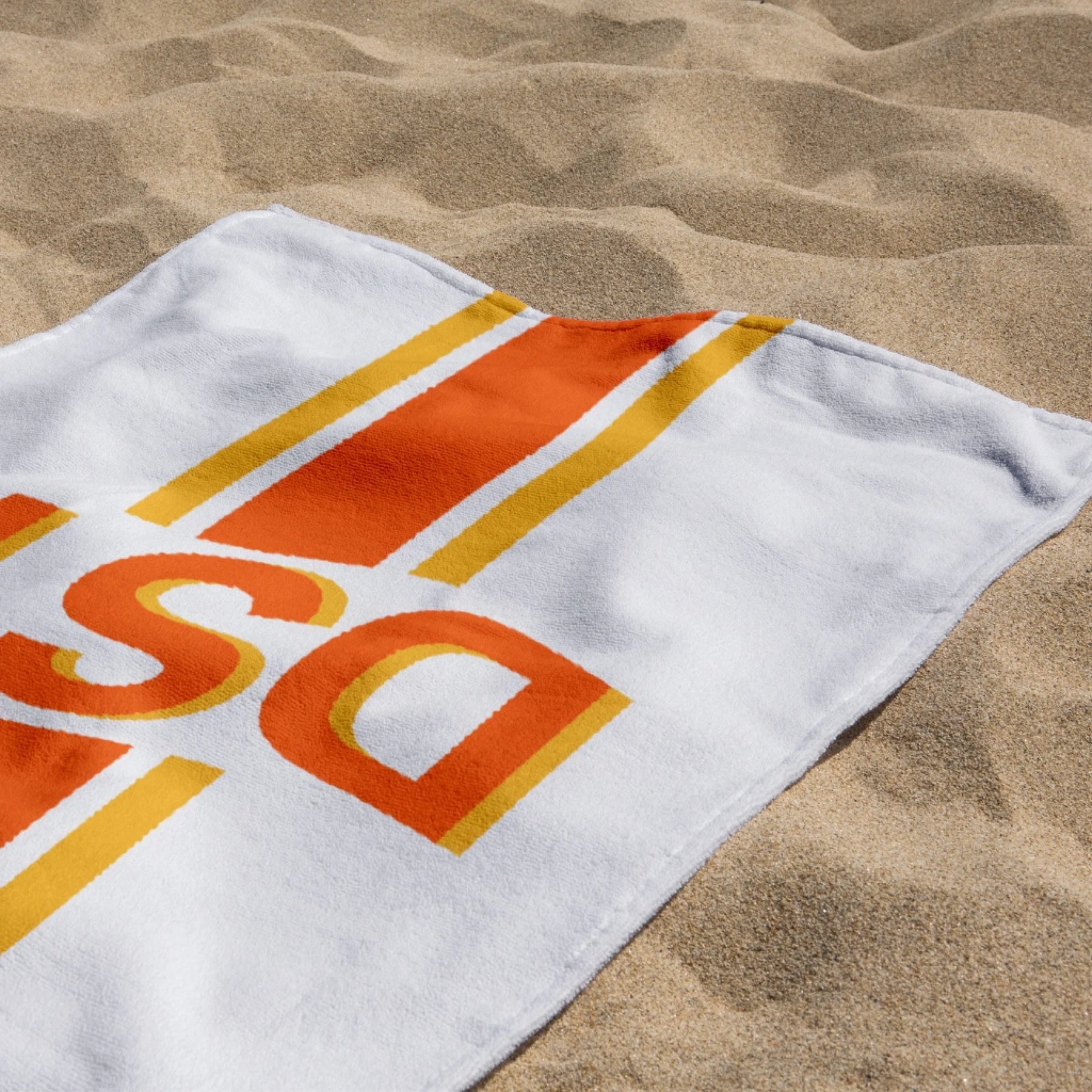 Close up of a personalized beach towel with orange DST monogram letters and colorful stripes, resting on sandy beach.