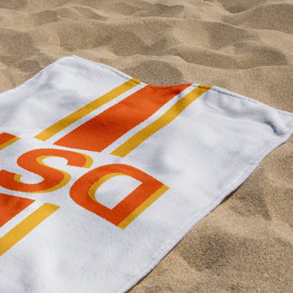 Close up of a personalized beach towel with orange DST monogram letters and colorful stripes, resting on sandy beach.