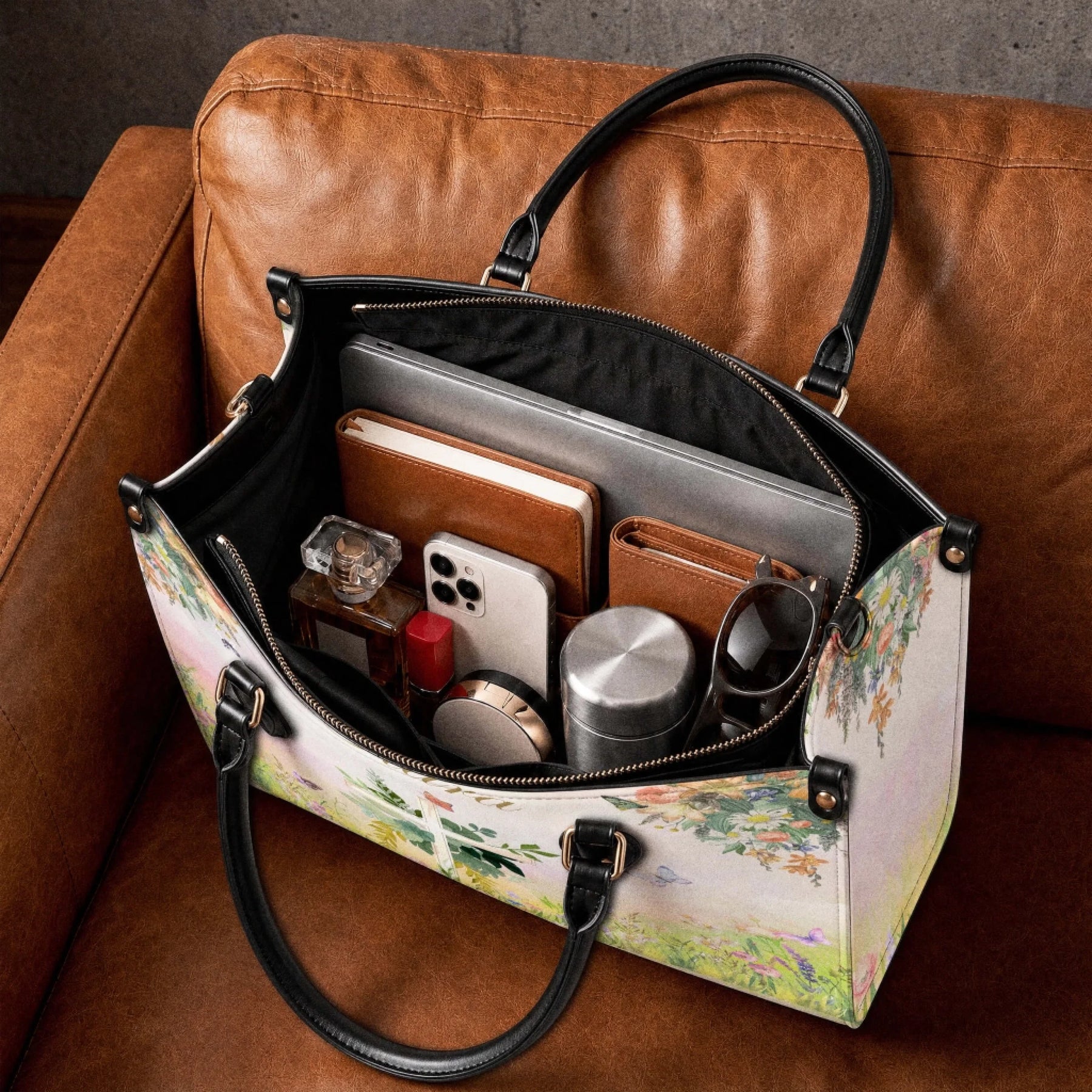 A personalized floral cross tote bag resting on a brown leather couch, filled with a laptop, wallet, phone, and perfume.