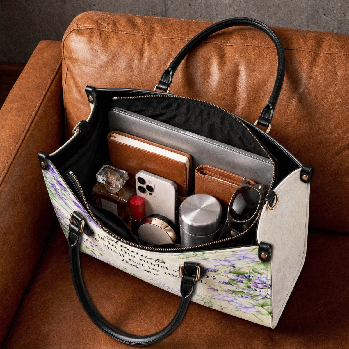 Spacious personalized floral handbag filled with daily items like a laptop, phone, perfume, and wallet on a couch.