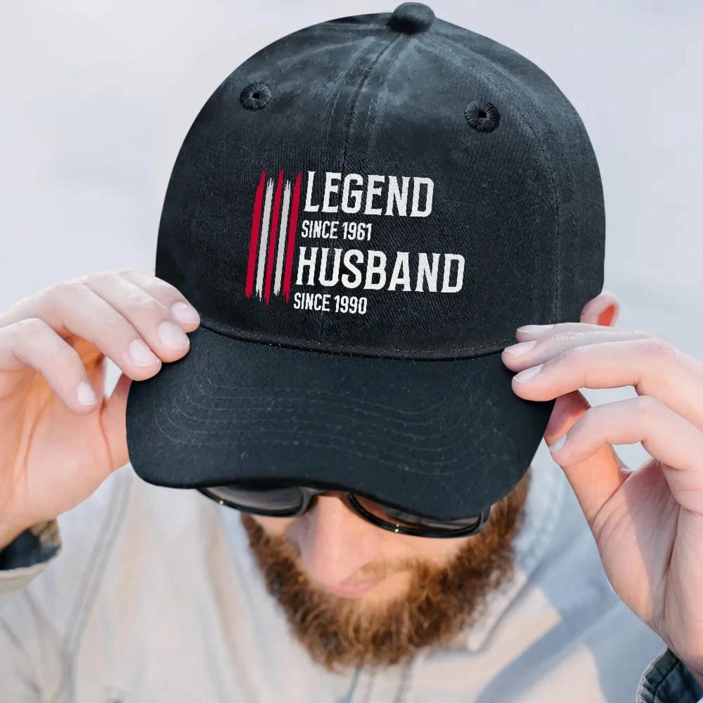Black cap with 'Legend Since 1961 Husband Since 1990' text held by a person.