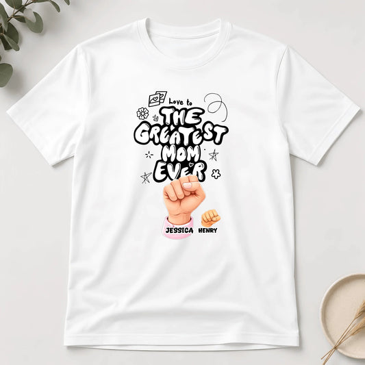 White t-shirt with 'The Greatest Mom Ever' design on a light gray background