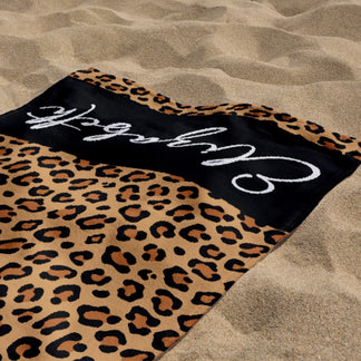 A customized leopard print beach towel with the name Elizabeth, lying flat on warm sand at the ocean shoreline