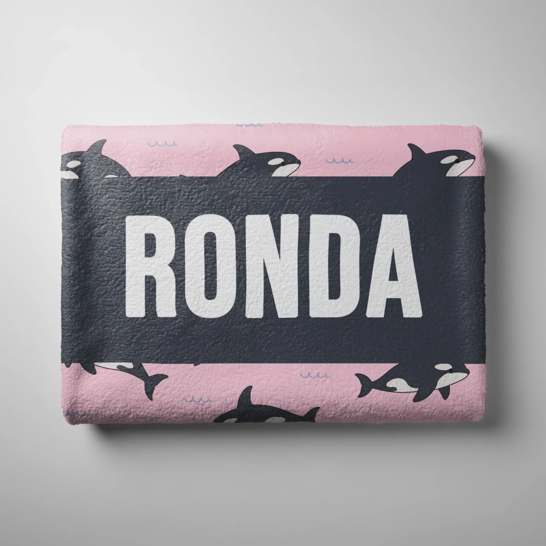 Folded pink beach towel featuring a repeating orca whale pattern and a dark center stripe personalized with the name Ronda.