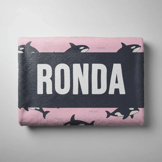 Folded pink beach towel featuring a repeating orca whale pattern and a dark center stripe personalized with the name Ronda.