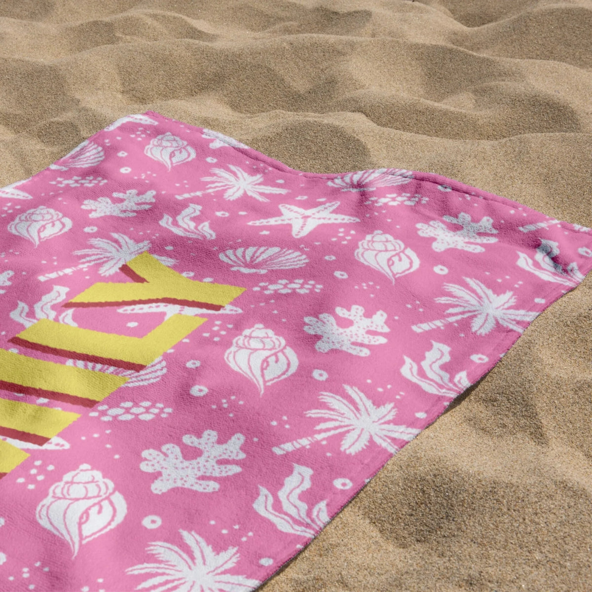Custom pink beach towel with a nautical seashell, palm tree, and coral pattern on sandy beach background.