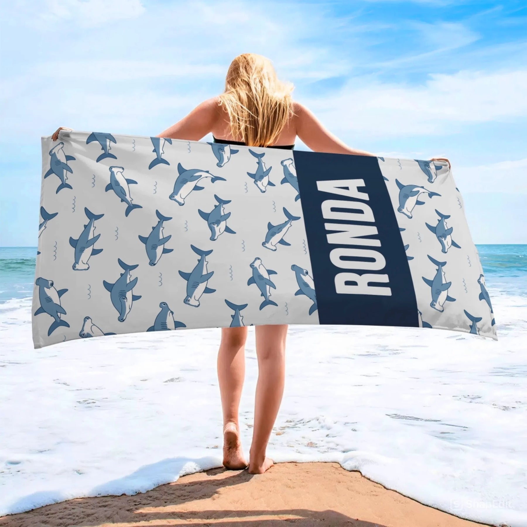 A woman holding a personalized beach towel with a shark pattern and the name Ronda printed on a dark blue background.