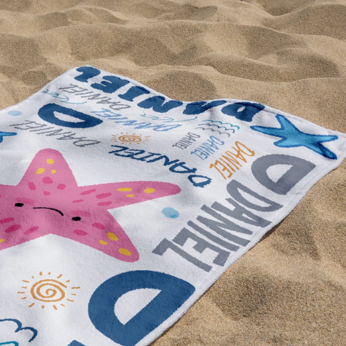 Folded custom beach towel with a friendly starfish pattern and the name Daniel resting on beach sand.