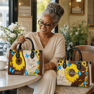 A woman holding personalized sunflower and butterfly tote bags featuring Be Still and Know That I Am God scripture.