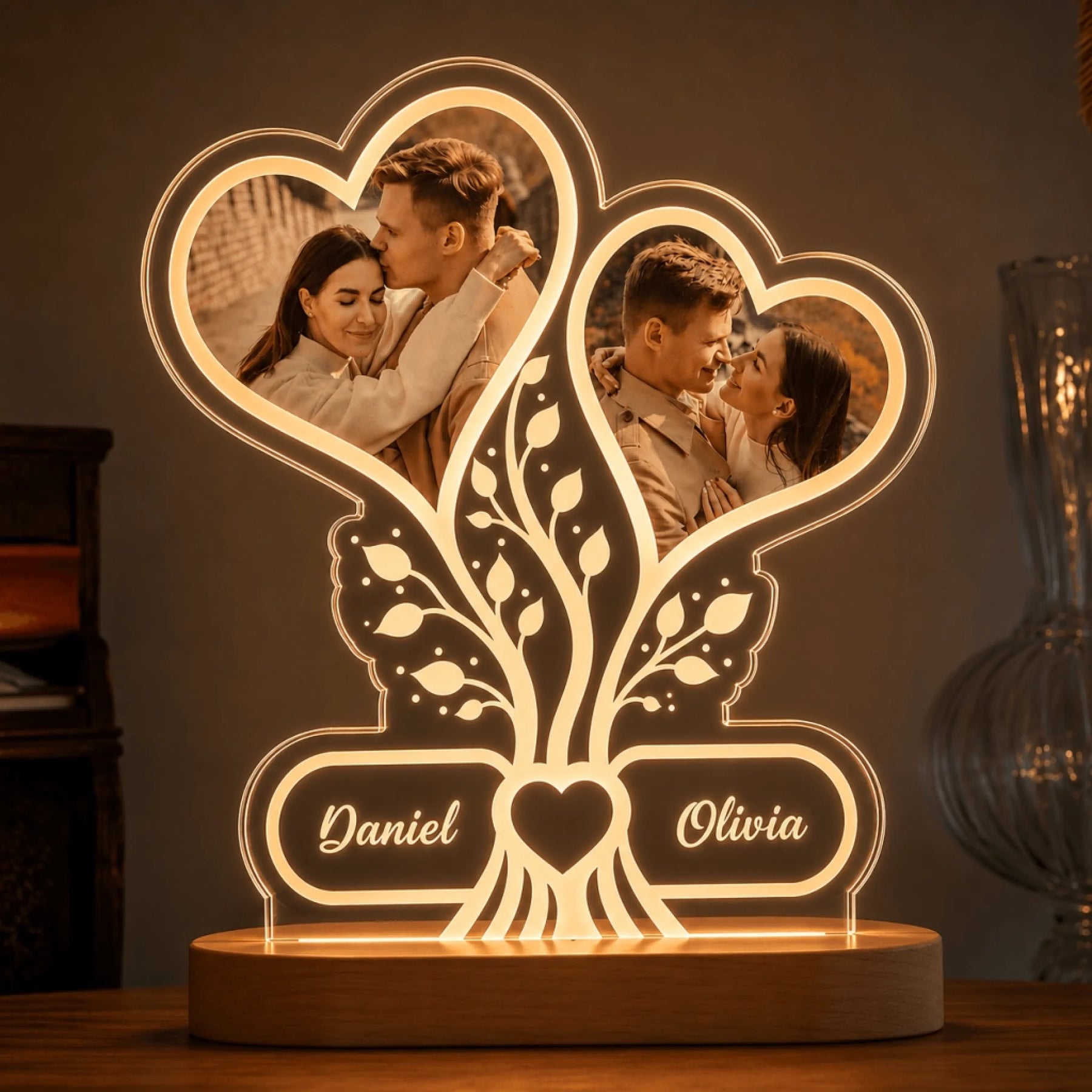 An illuminated acrylic desk lamp with a wooden base, featuring two heart-shaped photo frames containing images of a couple and a tree design with names.