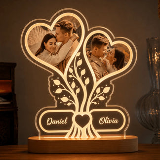An illuminated acrylic desk lamp with a wooden base, featuring two heart-shaped photo frames containing images of a couple and a tree design with names.