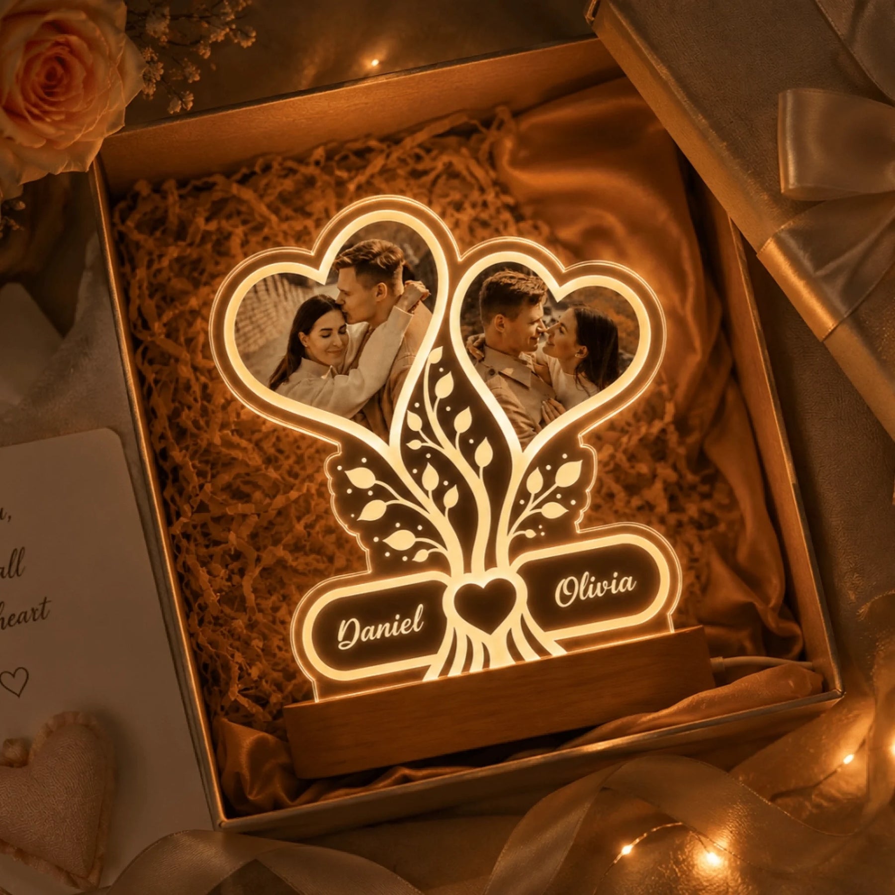 A glowing acrylic night light with two heart photo frames and a tree design, personalized with Daniel and Olivia, presented in an open gift box with shredded paper and a rose.