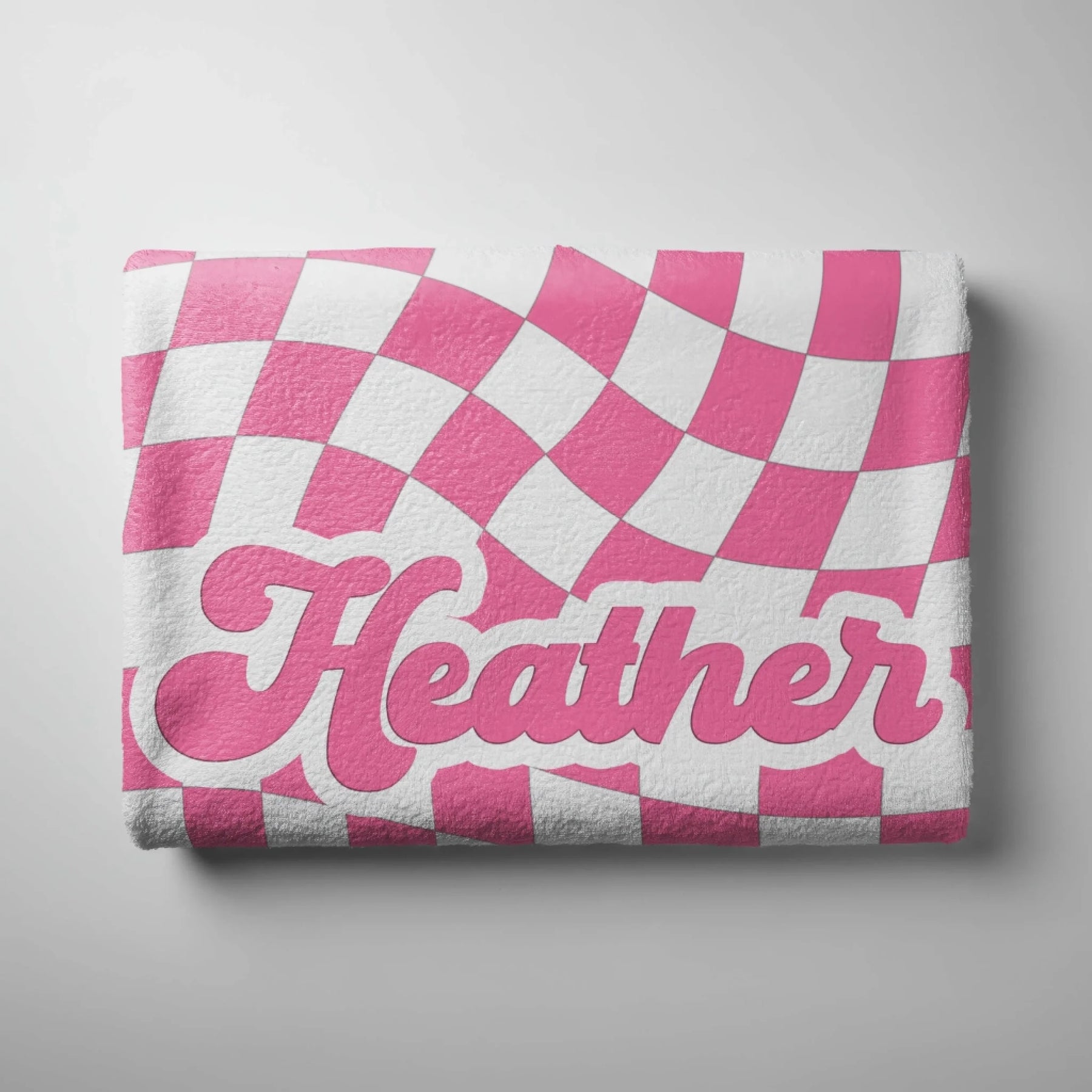 A folded beach towel showing a trendy pink and white wavy checkered pattern with the custom name Heather centered.