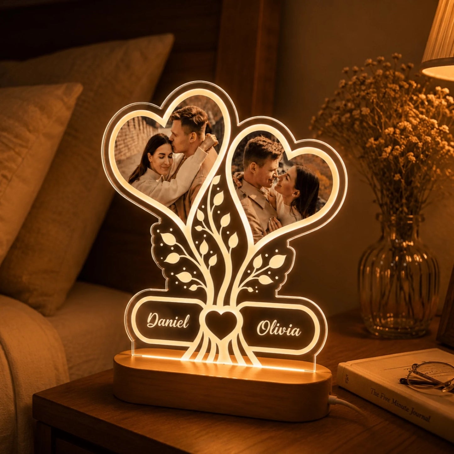 An illuminated acrylic night light featuring two heart photo frames with a couple, a tree design, and the names Daniel and Olivia, resting on a bedside table next to a book and glasses.
