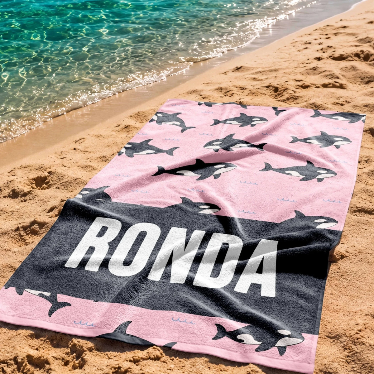Personalized pink beach towel decorated with an orca whale pattern and a bold black stripe with the name Ronda.