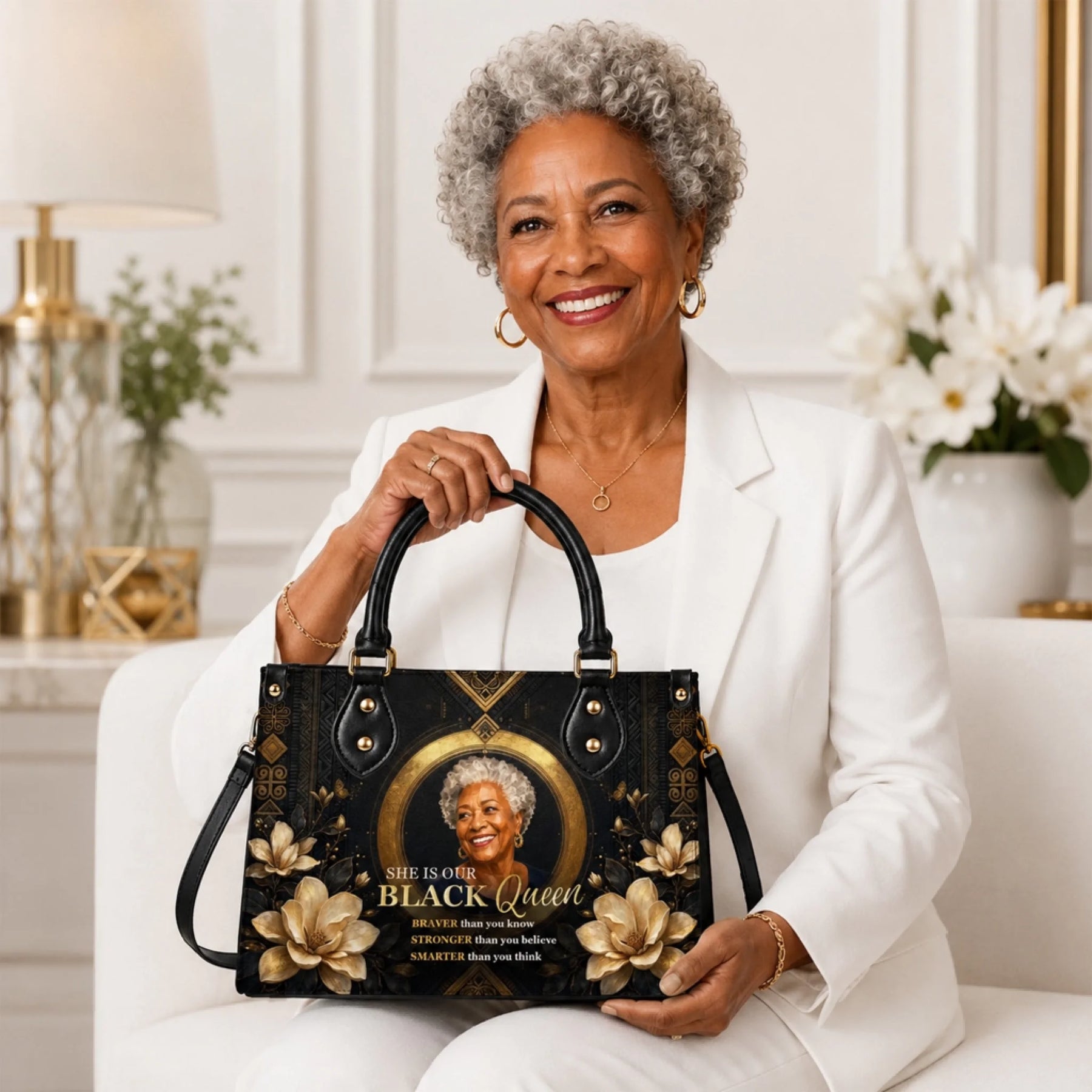 Smiling senior woman in a white suit holding a personalized black queen tote bag with gold floral accents and empowering text.