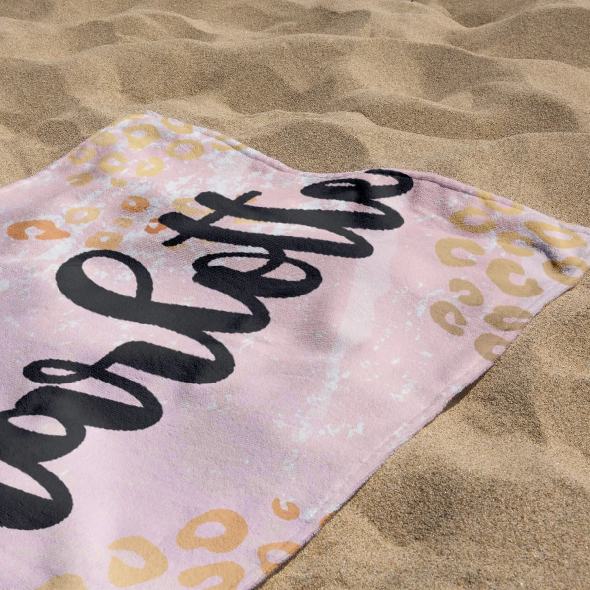 Close up of a personalized beach towel with leopard print pattern resting on beach sand on a sunny summer day.