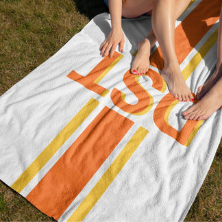 Top down view of a personalized DST monogram beach towel with orange stripes, used by a person lounging on the grass.
