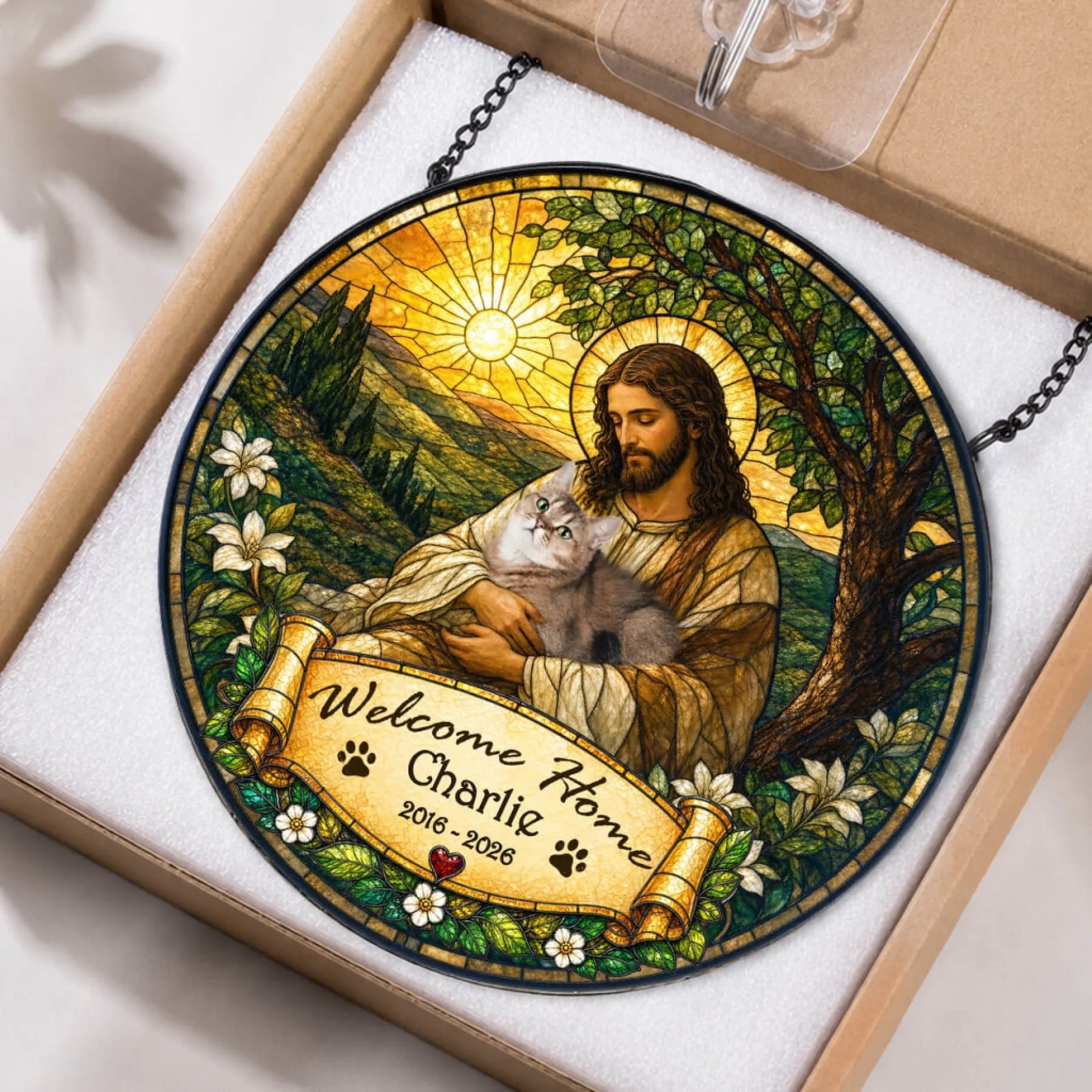 A round stained-glass style hanging with a robed figure holding a grey cat. A banner reads "Welcome Home Charlie" and "2016 - 2026". The item is nested in white foam inside an open cardboard box.