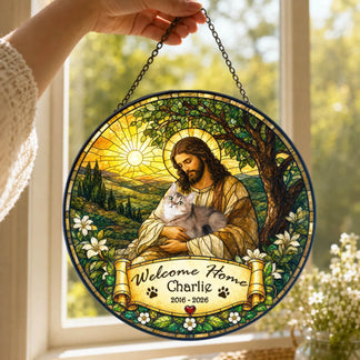 A hand holds up a round stained-glass style hanging. It features a robed figure with a halo holding a grey cat, with a banner reading "Welcome Home Charlie" and "2016 - 2026". It's seen against a bright window.