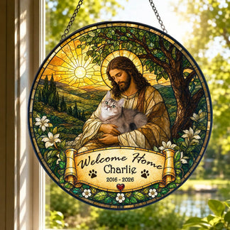 A round stained-glass style hanging depicting a robed figure with a halo holding a grey cat, against a sunny landscape. A banner reads "Welcome Home Charlie" and "2016 - 2026". It hangs in a bright window.
