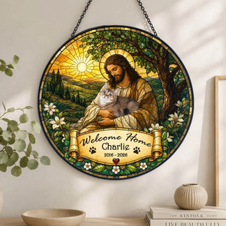 A round stained-glass style hanging with a robed figure holding a grey cat. A banner reads "Welcome Home Charlie" and "2016 - 2026". It's displayed on a shelf with plants and books.