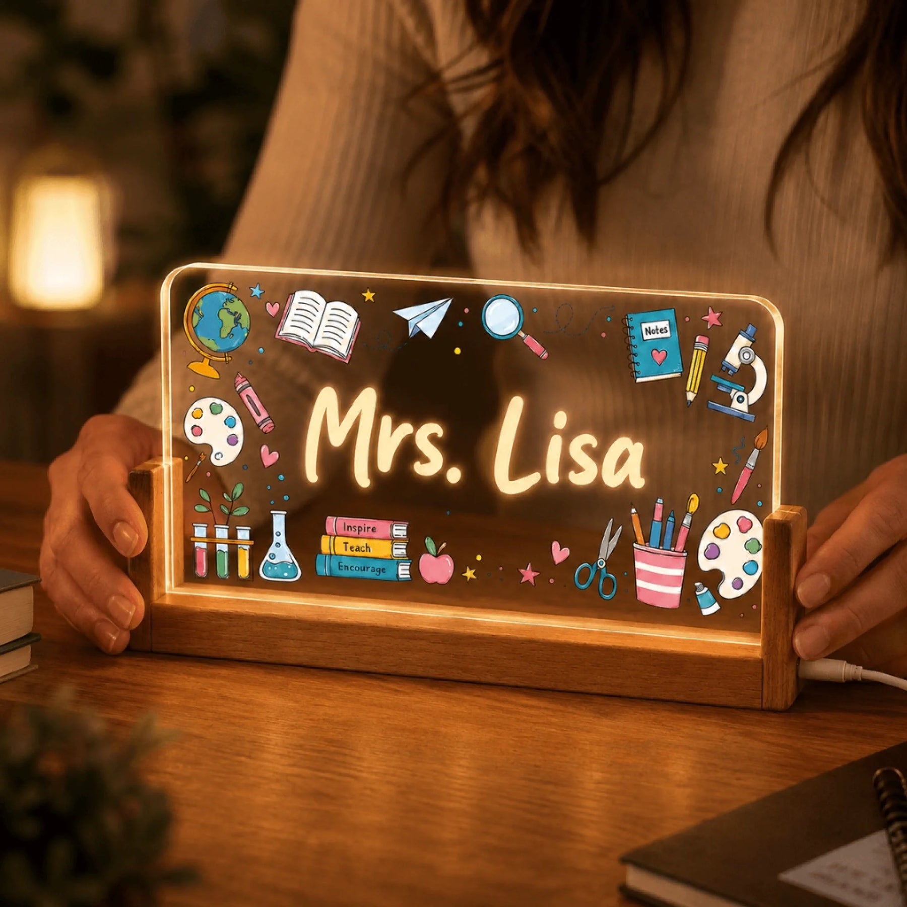 A person's hands holding an illuminated acrylic plaque with 'Mrs. Lisa' and colorful teaching illustrations, set in a wooden base on a desk.