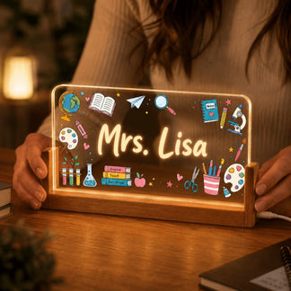 A person's hands holding an illuminated acrylic plaque with 'Mrs. Lisa' and colorful teaching illustrations, set in a wooden base on a desk.