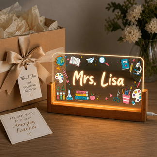 An illuminated personalized acrylic plaque for a teacher, featuring 'Mrs. Lisa', next to a gift box with a 'Thank You' tag and a greeting card.