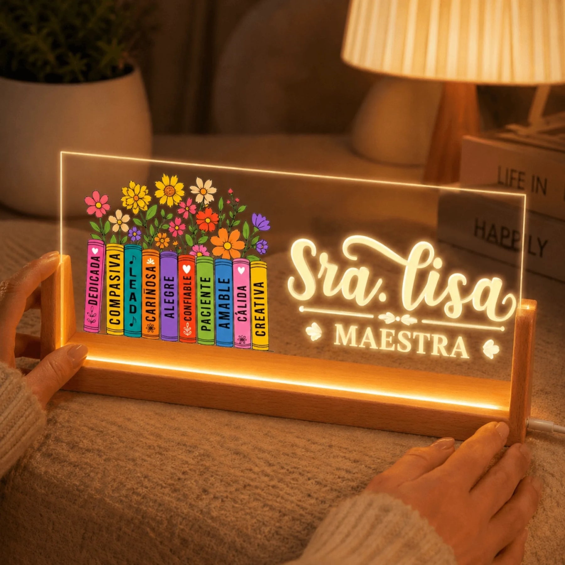 Two hands gently hold the wooden base of a lighted acrylic sign featuring colorful books and glowing text 'Sra. Lisa MAESTRA'.