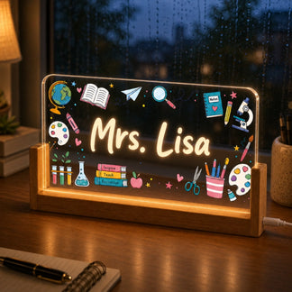 An illuminated personalized acrylic plaque for a teacher, featuring 'Mrs. Lisa' and various educational icons, on a desk in front of a rainy window.