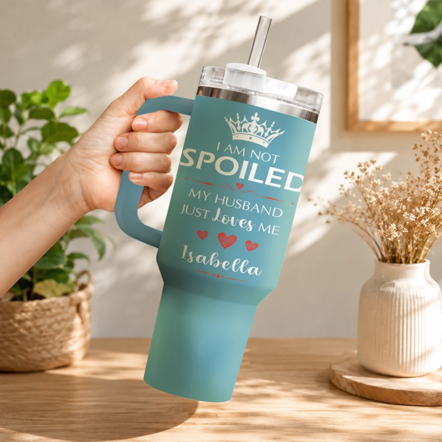 A hand holding a teal travel tumbler with I am not spoiled design against a home decor background.