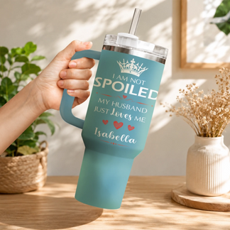 A hand holding a teal travel tumbler with I am not spoiled design against a home decor background.