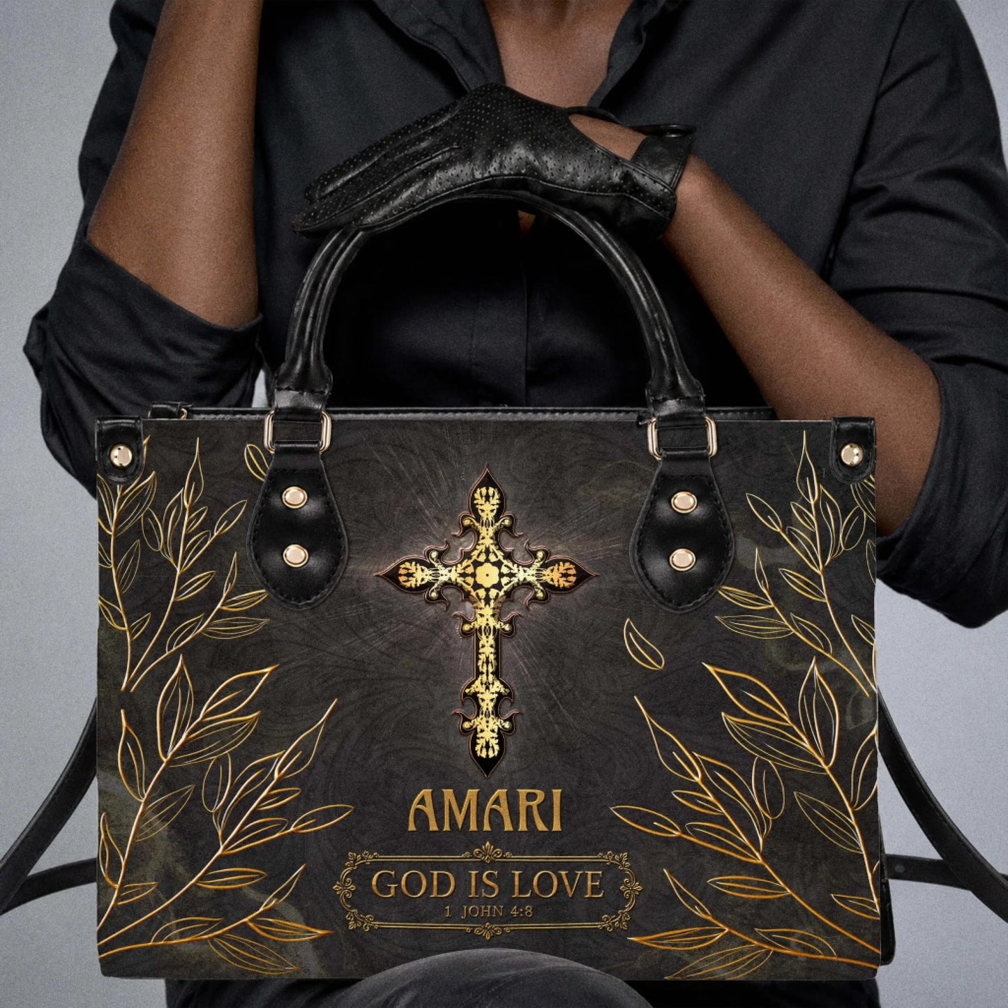 A person wearing black gloves holding a personalized black leather tote bag with a golden cross and biblical quote