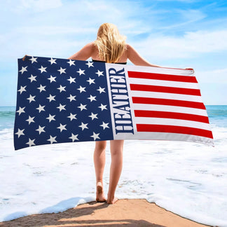A woman standing by the ocean holding a personalized American flag beach towel with the name Heather printed.