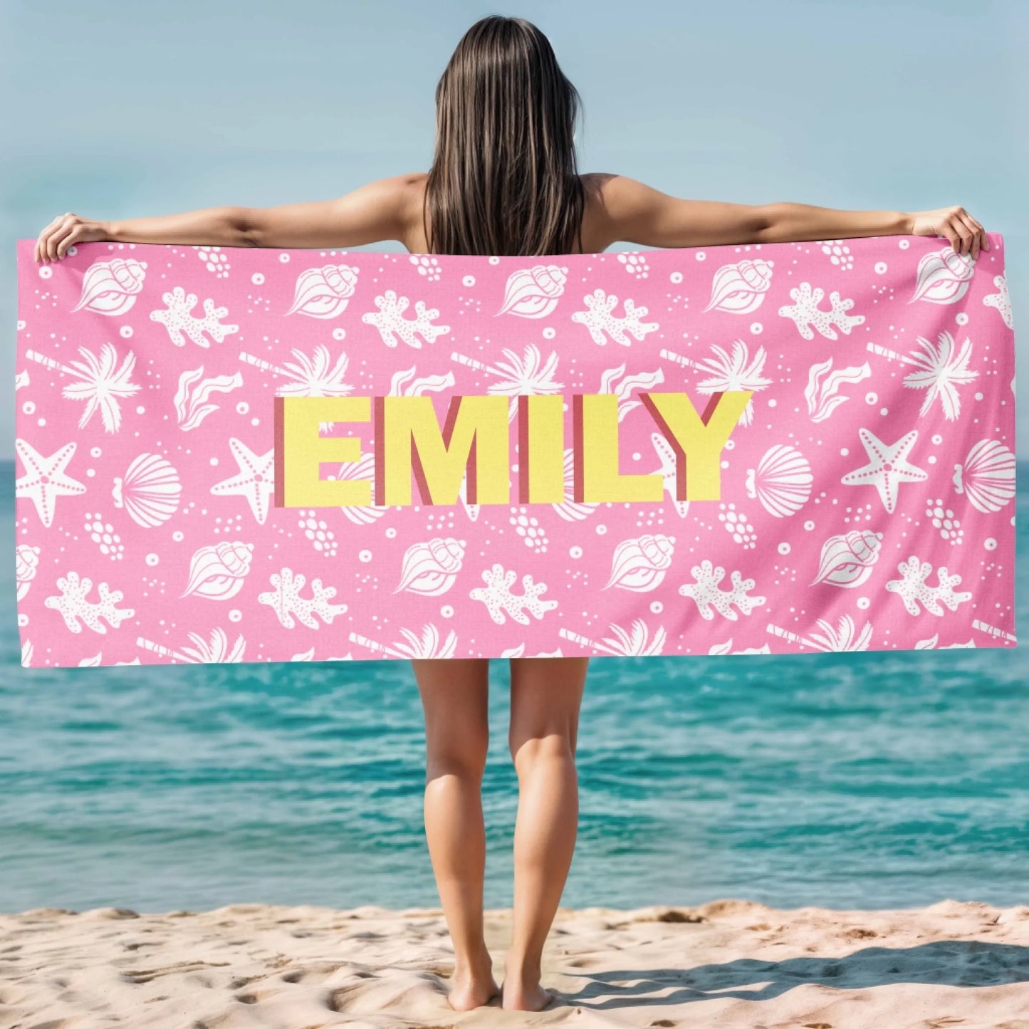 A woman at the beach holding a personalized pink beach towel with a seashell design and the custom name Emily on it.