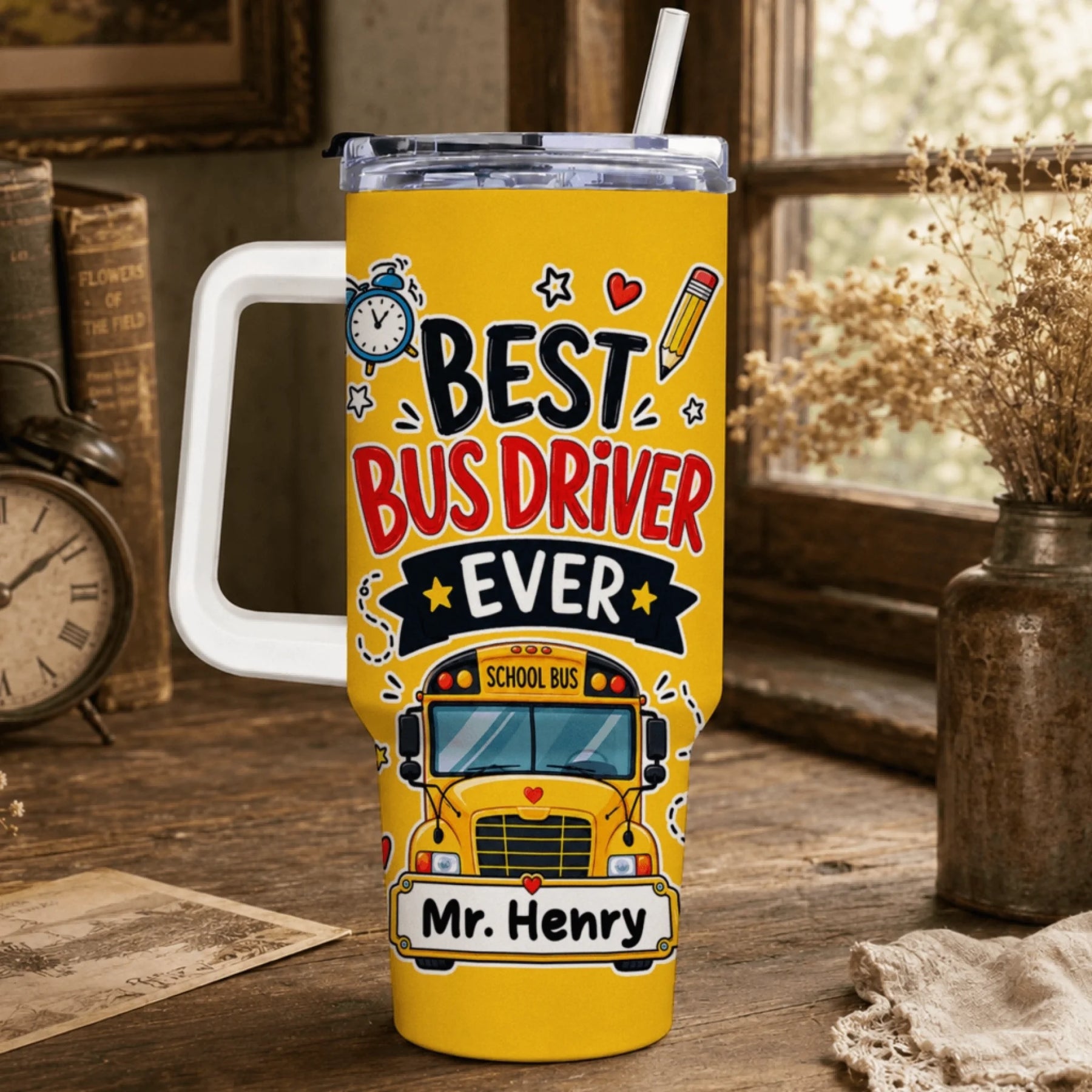 A yellow tumbler with a white handle and straw, displaying "BEST BUS DRIVER EVER", a school bus graphic, and "Mr. Henry" by a window.